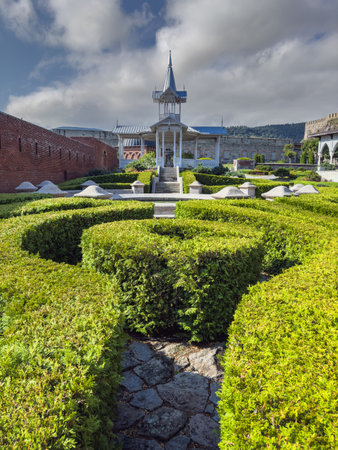 A picturesque garden with sculpted hedges, a pavilion, and a historic fortress with arched colonnades and stone walls in the background.の写真素材
