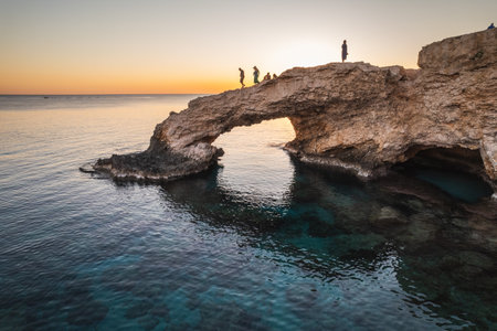 A breathtaking view of a natural rock arch over the sea, with a glowing sunset reflecting on the calm waters.の写真素材