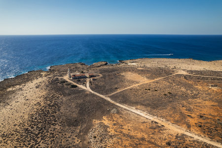 A remote lighthouse surrounded by rugged coastal terrain overlooking the vast, calm sea, captured from above.の写真素材