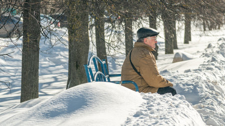 Senior man sitting on a bench in the snow in the park.の写真素材