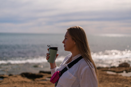 Young woman enjoying coffee from reusable cup while standing by the sea at sunset calm lifestyle...
