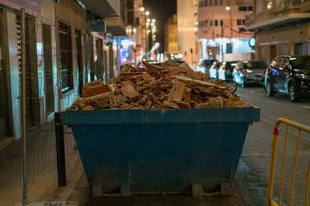 Blue dumpster filled with construction debris and broken bricks on urban street at night with parked...