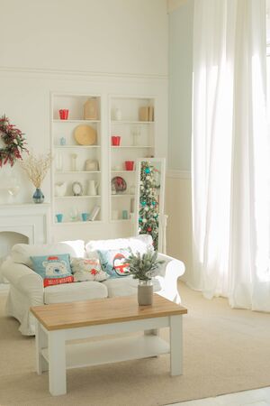 The interior of the Christmas room with a large window, a white sofa with pillows at the coffee...