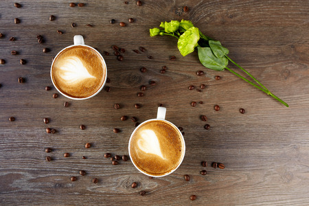 White cups of cappuccino on a wooden table with coffee beans and a green flowerの写真素材
