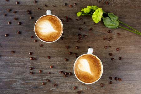 White cups of cappuccino on a wooden table with coffee beans and a green flowerの写真素材