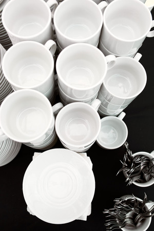 A table set of white mugs, plates and spoons on a buffet with a black tablecloth in a restaurantの写真素材
