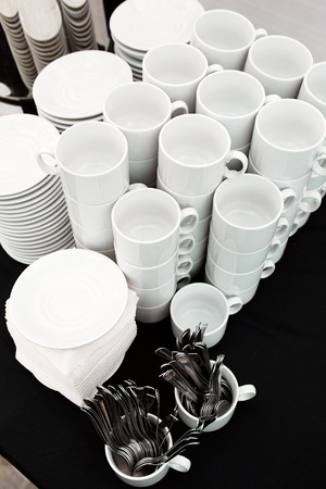 A table set of white mugs, plates and spoons on a buffet with a black tablecloth in a restaurantの写真素材