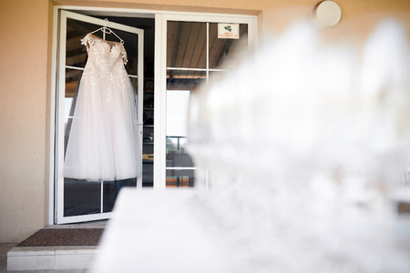 white wedding dress hanging on a white hanger on a white door with glasses of champagne in the backgroundの写真素材