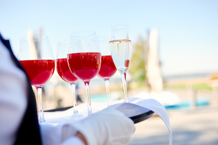 four wine glasses with red alcoholic drink and one glass of champagne on a tray of the waiter outdoorsの写真素材