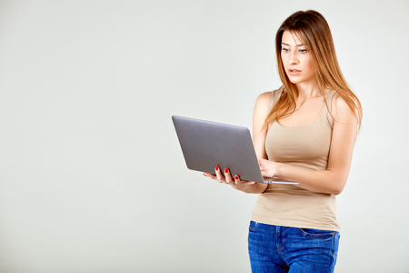 a girl in a beige t-shirt and blue jeans is standing against a gray wall holding a laptop and is looking at his screen in surpriseの写真素材