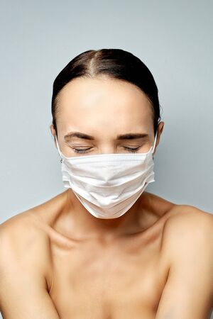 beautiful young brunette girl with pink manicure looks at a medical white disposable mask before putting on her on a gray background with emotions of fearの写真素材