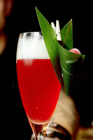 a beautiful red cocktail in a narrow glass with white froth and a green palm leaf decoration is held by a bartender in a light wooden bar counter against the backdrop of a barの写真素材