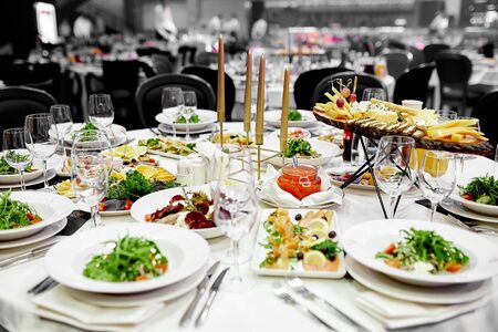 on the festive table with a white tablecloth on the table are brown festive candles and crystal wine glasses for drinks and plates with forks and knives for several people with a green vegetable salad and many snacksの写真素材
