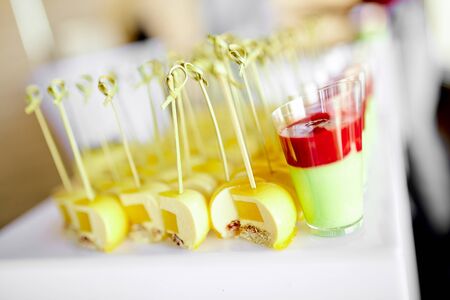 Catering, delicious new dessert canapes with yellow mango on a skewer and red-green mousse dessert in a glass of wine stand next to the table for treating guests of the eventの写真素材
