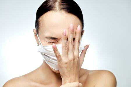 A girl in a medical mask on her face with possible signs of a coronavirus in fear holds her face in her hand with a pink manicure on a gray backgroundの写真素材