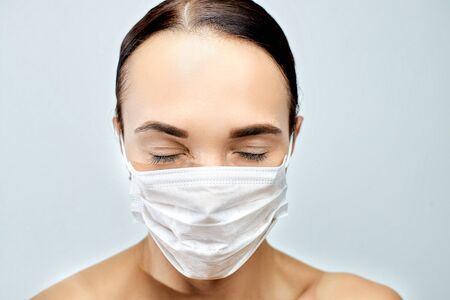 A brunette girl with closed eyes and long eyelashes holds in both hands with a manicure a white medical mask on a gray backgroundの写真素材