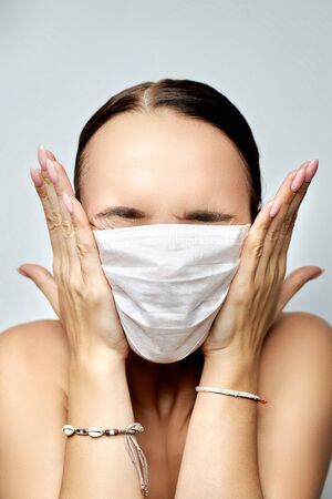 beautiful young brunette girl with pink manicure looks at a medical white disposable mask before putting on her on a gray backgroundの写真素材