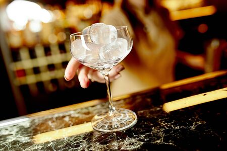 a girl holds a crystal glass with ice on a marble bar counter against the background of a bar and warm lightの写真素材