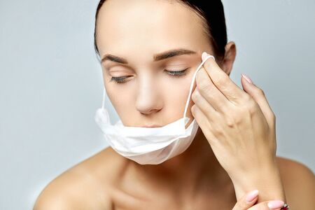 young brunette girl looking down with a tired look with a flat white medical disposable mask on a long neck near the collarbone and well-groomed hands with pink manicure on a gray backgroundの写真素材