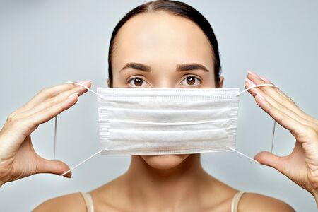 beautiful brunette girl puts on a medical mask on a gray background with pink manicure and a neat hairstyle with a confident lookの写真素材