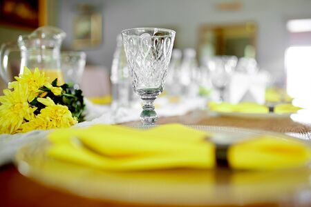 on the table there is a crystal empty wine glass, in front there is a round transparent plate on which there are appliances and covered with a yellow festive napkin, next to them are yellow flowersの写真素材
