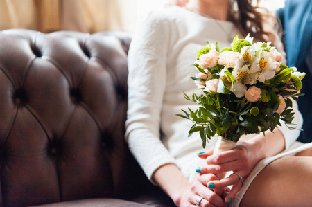 Bride and groom holding wedding bouquet together . Hug . He is sitting on a leather couchの写真素材