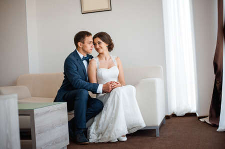 Wedding couple indoors is hugging each other. Beautiful model girl in white dress. Man in suit. Beauty bride with groom. Female and male portrait. Woman with lace veil. Cute lady and handsome guyの写真素材
