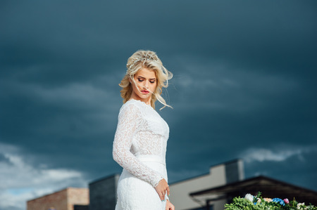 Beautiful young bride woman in luxury wedding dress with red lips makeup and long wavy hairstyle. Beauty girl portrait posing against the background of dark rainy clouds, outdoor photo.の写真素材