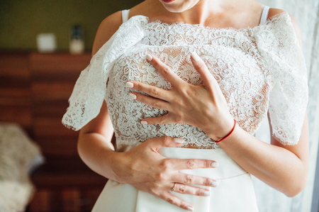 Wedding. Bride. Preparations. Wedding Dress. Bride in cream-colored lace dress stands in front of the window with her hands foldedの写真素材