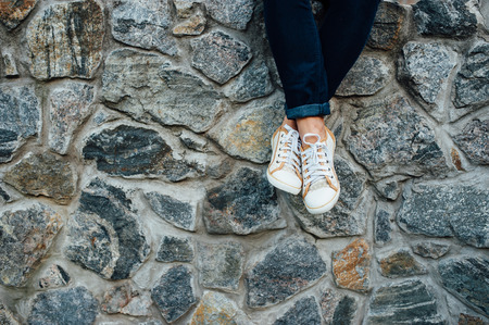 Young fashion man's legs with sneakers seated in stone wall. White shoes