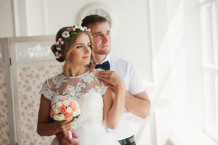 Wedding couple indoors is hugging each other. Beautiful model girl in white dress. Man in suit. Beauty bride with groom. Female and male portrait. Woman in beautiful wreath of flowers.の写真素材
