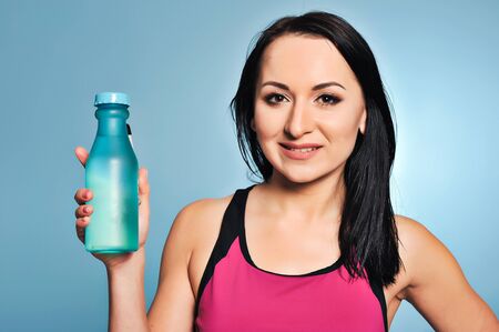 Cheerful charming fitness girl in pink top showing a bottle of water over blue backgroundの写真素材
