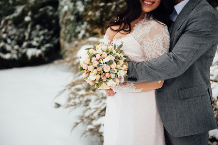 Beautiful wedding bouquet in hands of the bride with the big beautiful breasts. Image taken in the street in winter. the groom gently embraces the brideの写真素材