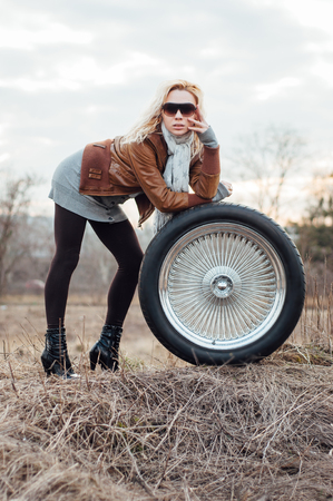 Sexy young blonde girl sits on a large-sized wheel with thin knitting needles. The model is dressed in leggings and a brown jacketの写真素材