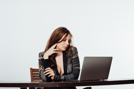 Sexy business woman in a jacket with a big neckline and chest sits on a chair and works with a laptopの写真素材