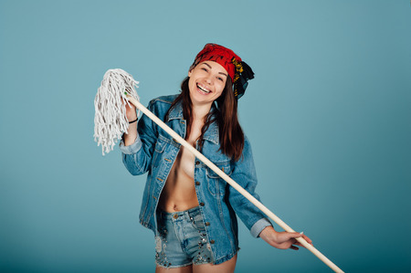 Beautiful sexy girl with a mop in her hands is wearing jeans clothes. She stands on blue backgroundの写真素材