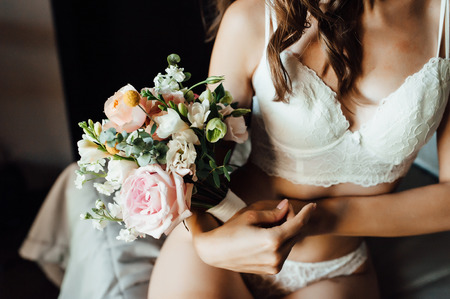 Sexy beautiful nude brunette bride with bouquet and bright smoky eyes make up and wavy hair style in white erotic lingerie underwear in sunny room of the hotel. Morning of the bride at wedding dayの写真素材