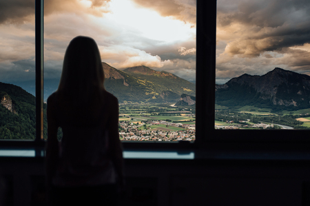 silhouette of woman in sunshine at window with view on sunset in mountainsの写真素材