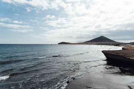 Many colorful kites on beach and kite surfers riding waves during windy day in canarian El Medano in Tenerife with Montana roja hills on horizonの写真素材