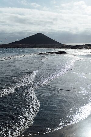 Many colorful kites on beach and kite surfers riding waves during windy day in canarian El Medano in Tenerife with Montana roja hills on horizonの写真素材