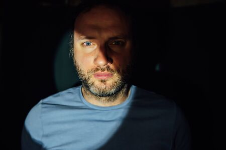 Portrait of young man on black background with hard light. He has a serious expression on the face like thinking or meditating. He has short hair but natural and a little beard and brown eyes.の写真素材