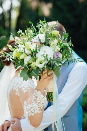 Cheerful newlyweds are hugging at their wedding and covering their face with a wedding bouquetの写真素材
