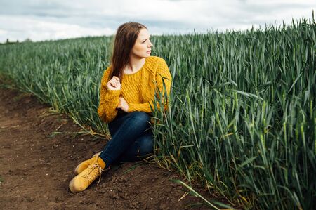 Beautiful girl with jeans, a yellow sweater and boots sits near an early wheat fieldの写真素材