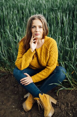 Beautiful girl with jeans, a yellow sweater and boots sits near an early wheat fieldの写真素材