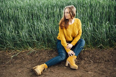 Beautiful girl with jeans, a yellow sweater and boots sits near an early wheat fieldの写真素材