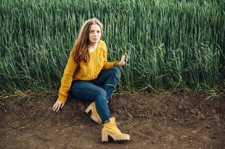 Beautiful girl with jeans, a yellow sweater and boots sits near an early wheat fieldの写真素材