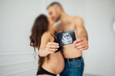 A happy married couple is holding in their hands a snapshot of an ultrasonic study of the baby. Husband hugs a beautiful adult pregnant wife. Second trimesterの写真素材