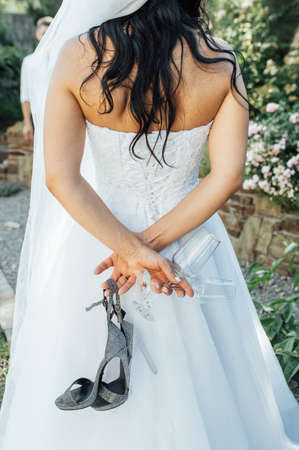 Beautiful bride stands with her back and holds her shoes and a glass of champagneの写真素材