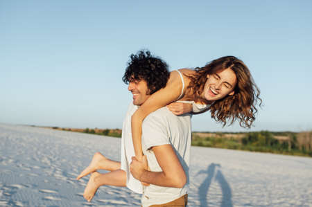 Beautiful couple having fun on the beach. Cheerful girl jumped on her boyfriends backの写真素材