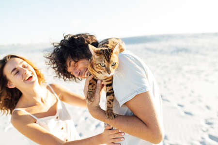 Cheerful young couple having fun on the beach with their bengal catの写真素材
